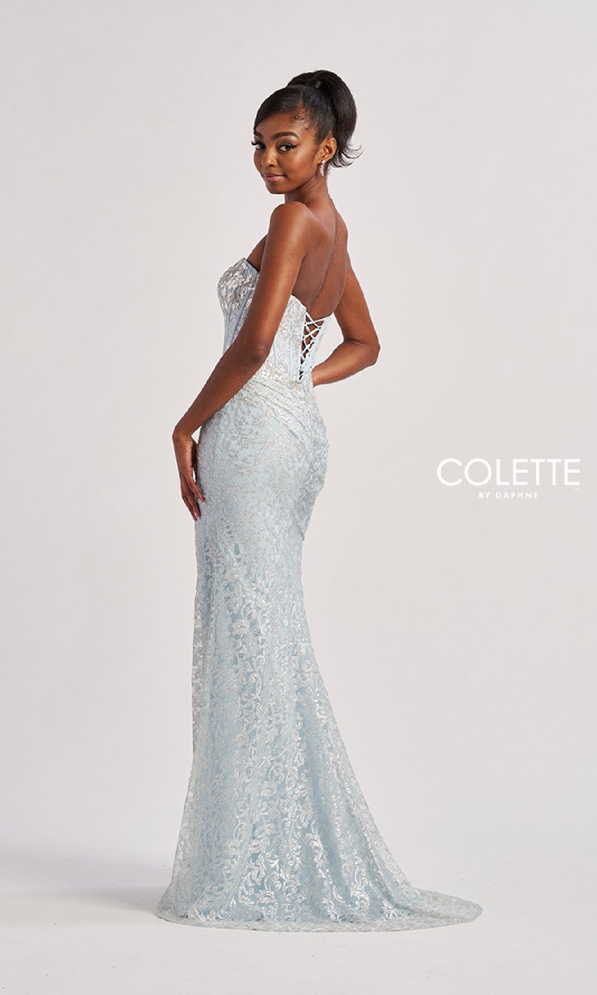 A woman stands in a studio wearing the Colette CL8595 Long Prom Dress—a light blue, strapless, floor-length lace gown with a lace-up back. The